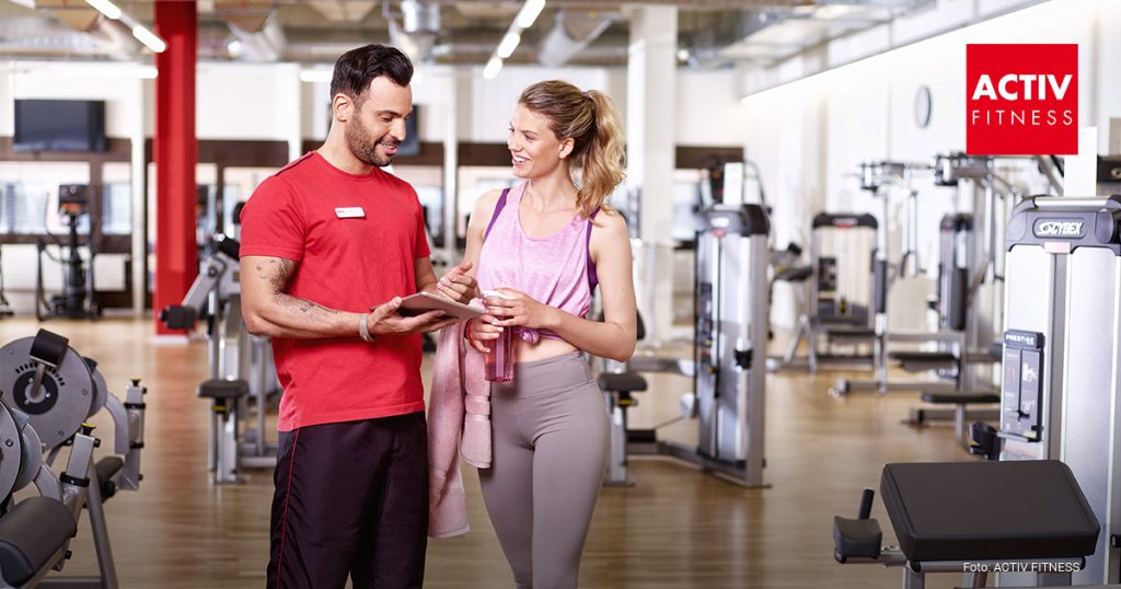 Ein Trainer steht im Fitnessstudio nebem einer Frau die dort trainiert und erzählt ihr etwas