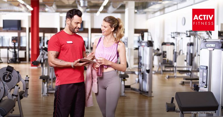 Ein Trainer steht im Fitnessstudio nebem einer Frau die dort trainiert und erzählt ihr etwas